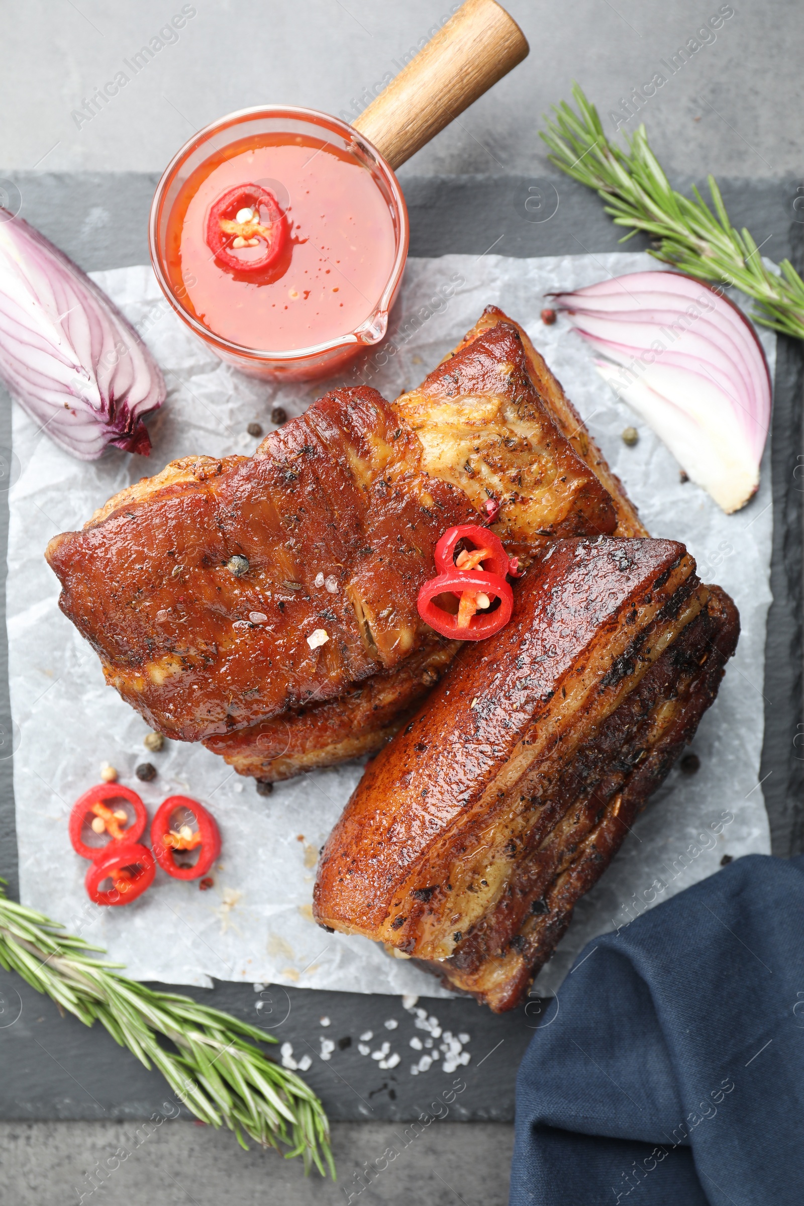 Photo of Pieces of baked pork belly served with sauce, rosemary and chili pepper on grey table, top view