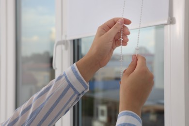 Photo of Woman opening white roller blinds on window indoors, closeup