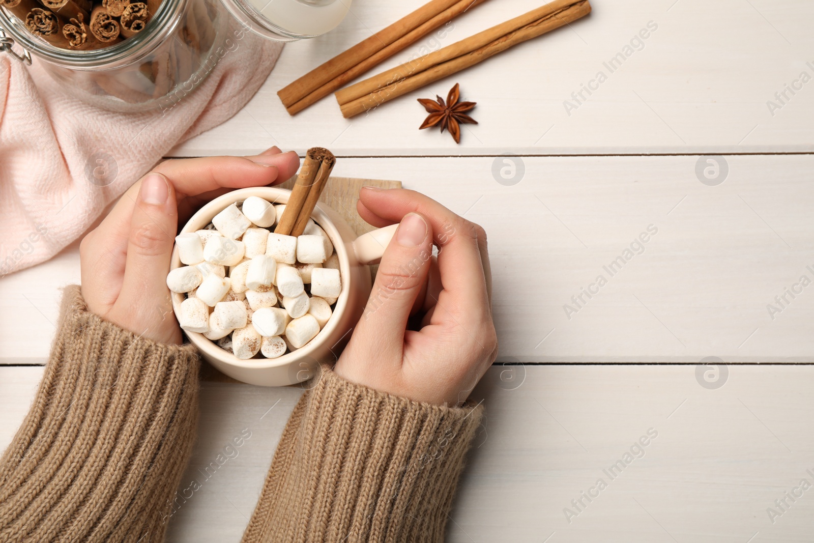 Photo of Woman holding cup of hot drink with marshmallows and aromatic cinnamon at white wooden table, top view. Space for text