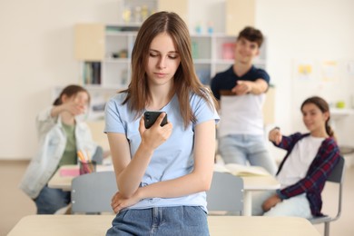 Photo of Teen problems. Lonely girl with smartphone standing separately from other students in classroom