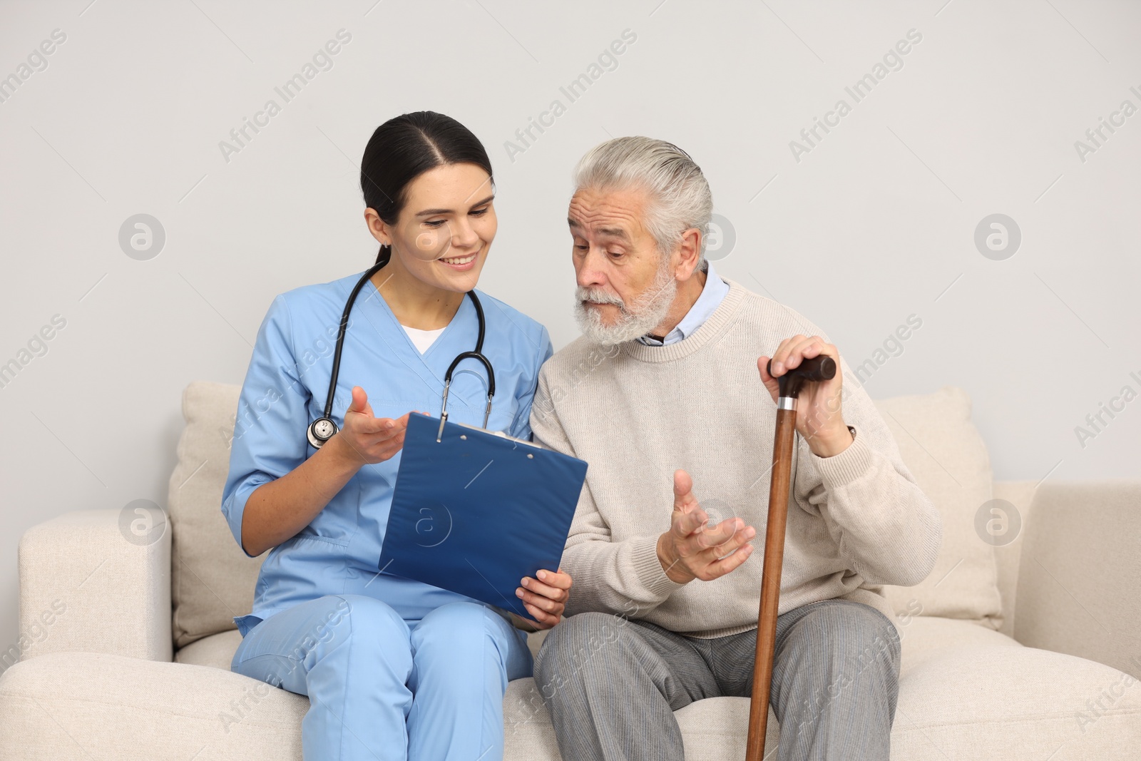 Photo of Smiling nurse with clipboard assisting elderly patient on sofa indoors