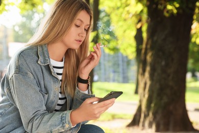 Photo of Young woman smoking cigarette while using smartphone outdoors, space for text
