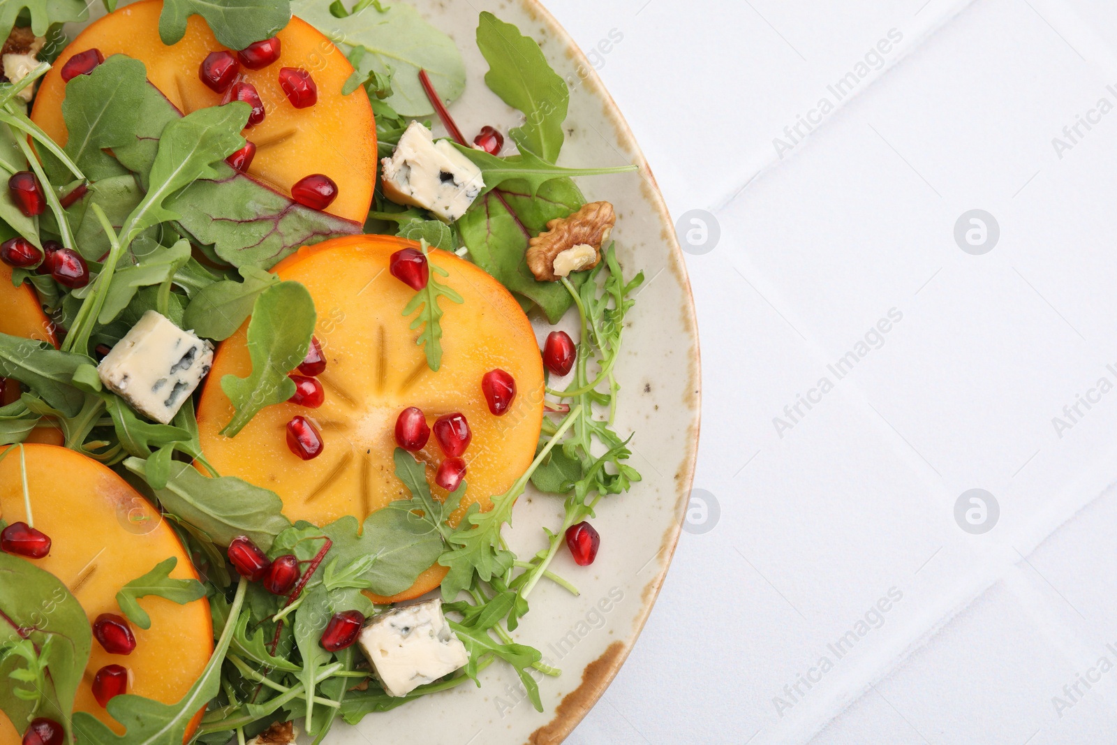 Photo of Tasty salad with persimmon, blue cheese, pomegranate and walnuts served on white tiled table, top view. Space for text