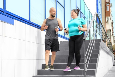 Photo of Overweight man and woman running with dumbbells on stairs outdoors