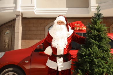 Photo of Authentic Santa Claus near car with fir tree and bag full of presents outdoors
