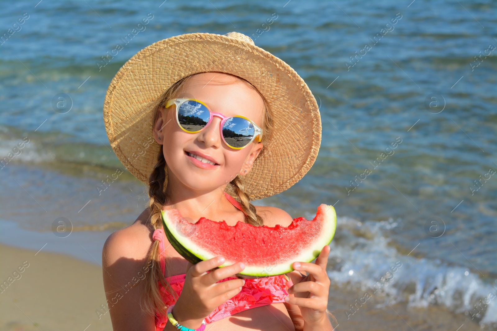 Photo of Cute little girl in straw hat and sunglasses eating juicy watermelon on beach