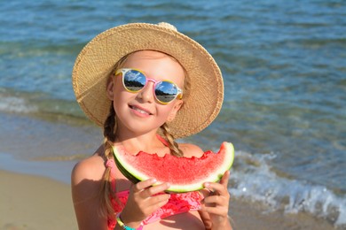 Photo of Cute little girl in straw hat and sunglasses eating juicy watermelon on beach