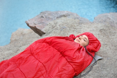 Image of Young camper resting in sleeping bag on cliff near lake