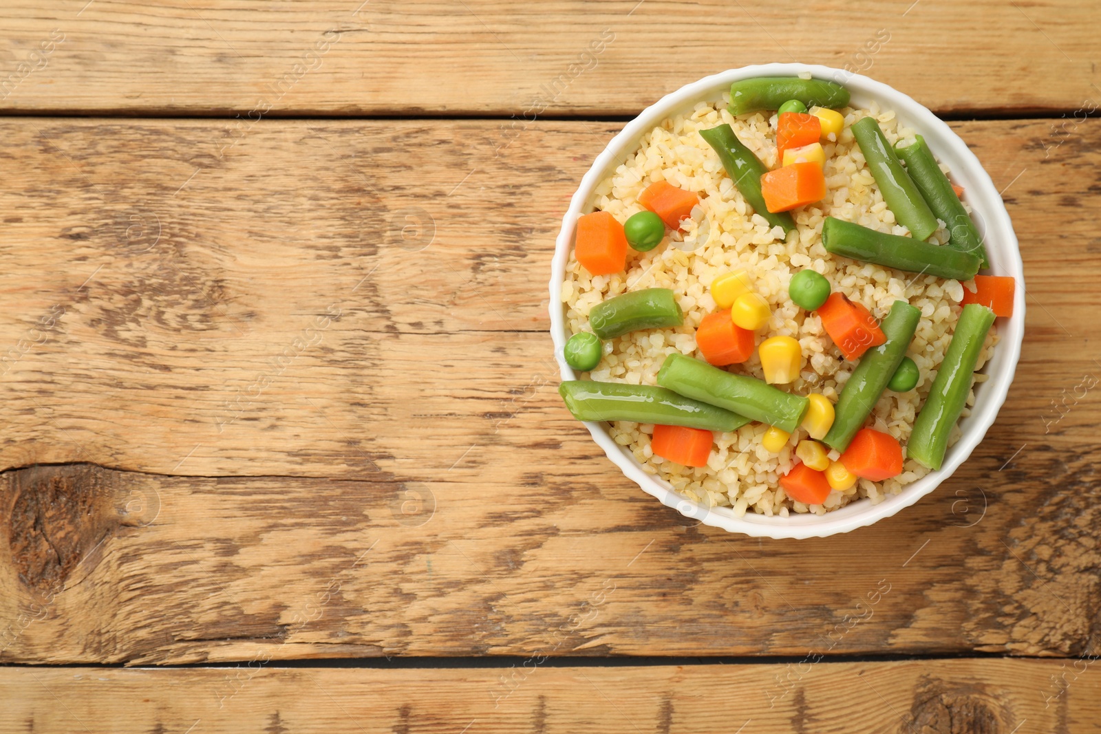 Photo of Delicious bulgur with vegetables in bowl on wooden table, top view. Space for text