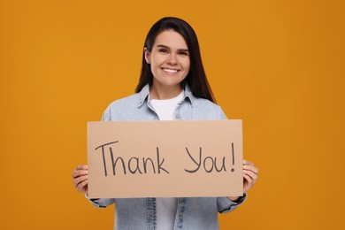 Photo of Happy woman holding cardboard sheet with phrase Thank You on orange background