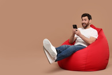 Photo of Happy man with smartphone sitting on bean bag against light brown background. Space for text