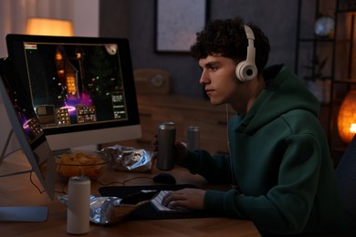 Young man with energy drink and headphones playing video game at wooden desk indoors