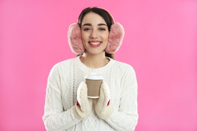 Beautiful young woman in earmuffs with cup of drink on pink background