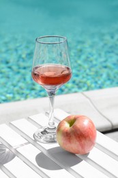 Photo of Glass of tasty rose wine and apple on wooden table near swimming pool