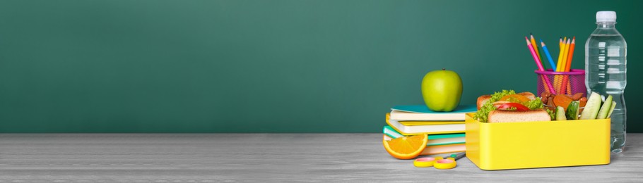 Image of School meal. Lunch box with healthy food and different stationery on wooden table near green chalkboard, space for text. Banner design