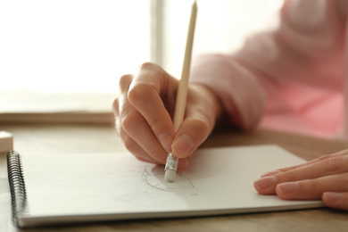 Photo of Woman correcting picture in notepad with pencil eraser at wooden table, closeup