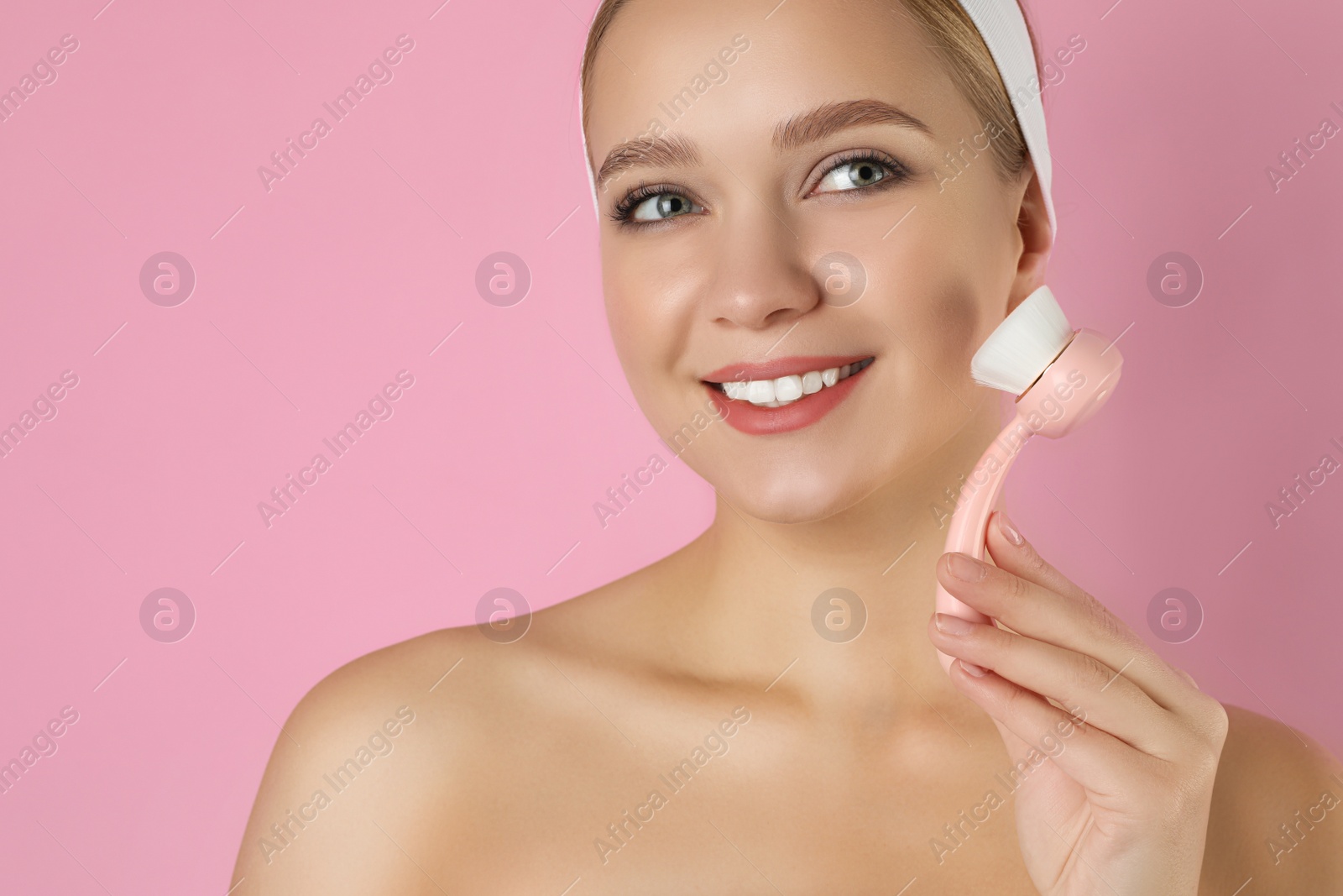 Photo of Young woman washing face with cleansing brush on pink background. Cosmetic product