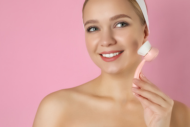 Photo of Young woman washing face with cleansing brush on pink background. Cosmetic product