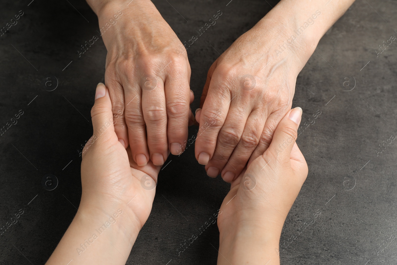 Photo of Young and elderly women holding hands at dark grey table, closeup