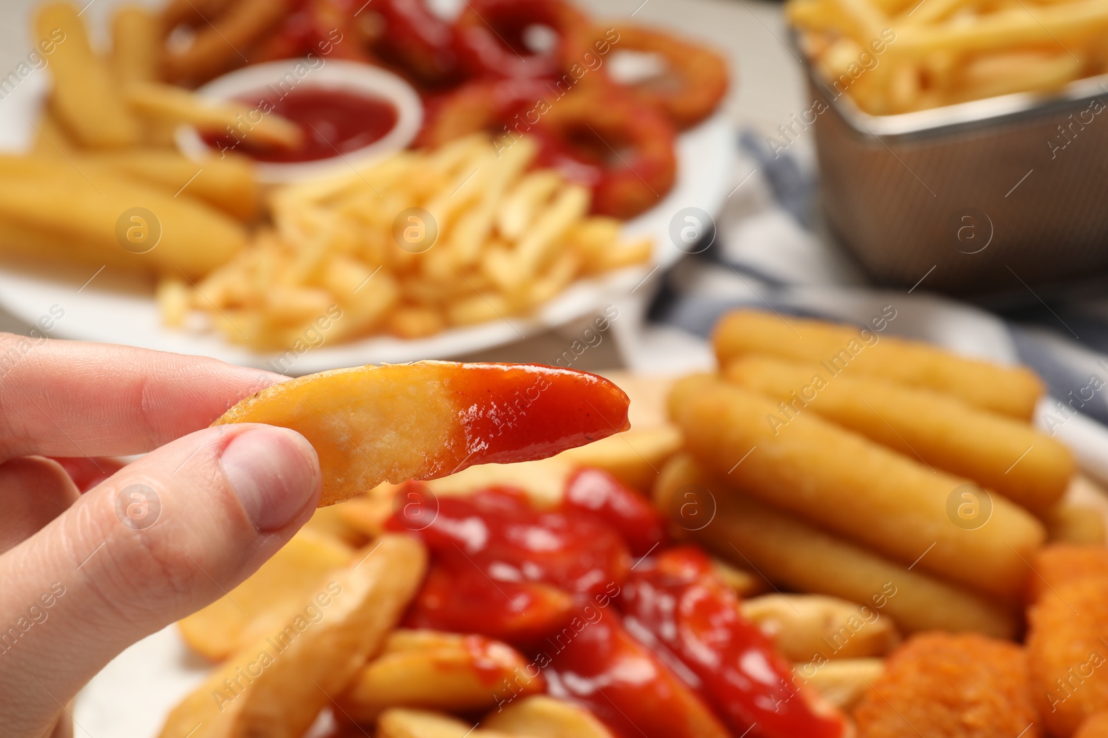 Photo of Woman holding delicious baked potato wedge with ketchup near different snacks at table, closeup