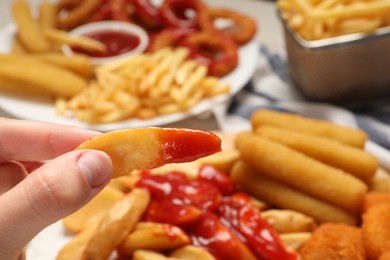 Photo of Woman holding delicious baked potato wedge with ketchup near different snacks at table, closeup