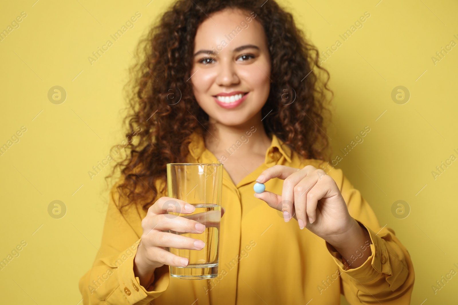 Photo of African-American woman with glass of water and vitamin pill against yellow background, focus on hands