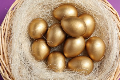 Photo of Shiny golden eggs in nest on violet background, top view