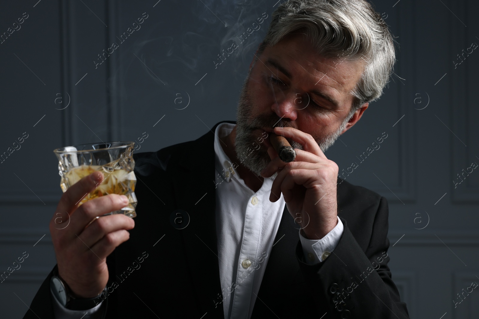 Photo of Handsome bearded man with glass of whiskey smoking cigar against dark grey background