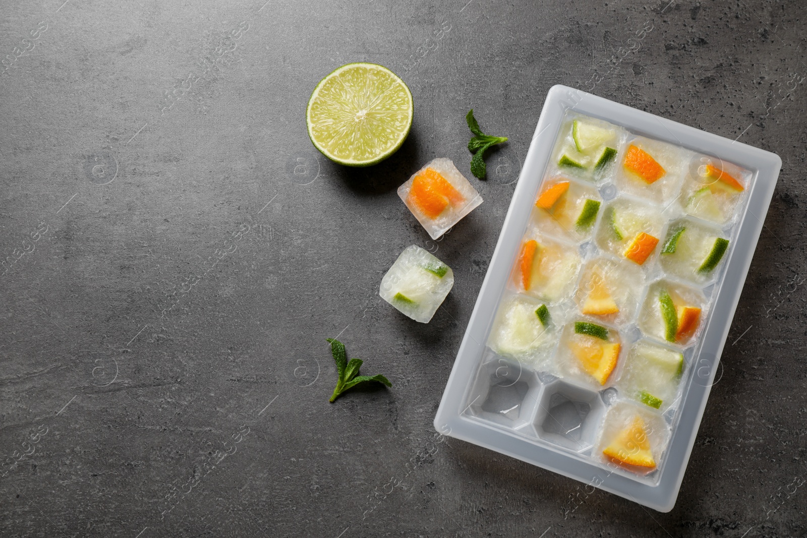 Photo of Ice cube tray with frozen fruits and mint on grey table, flat lay. Space for text