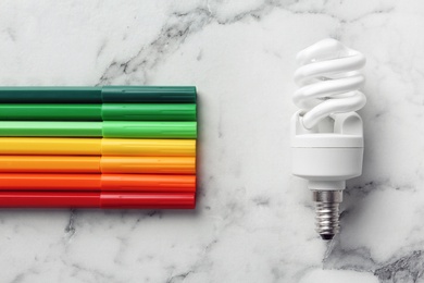 Photo of Flat lay composition with colorful markers and fluorescent light bulb on white marble background. Energy efficiency concept