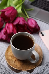 Photo of Beautiful tulips, cup of coffee, laptop, smartphone and sweater on light gray blanket, closeup