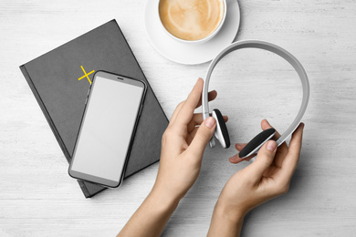 Photo of Woman with Bible, phone, cup of coffee and headphones at white wooden table, top view. Religious audiobook