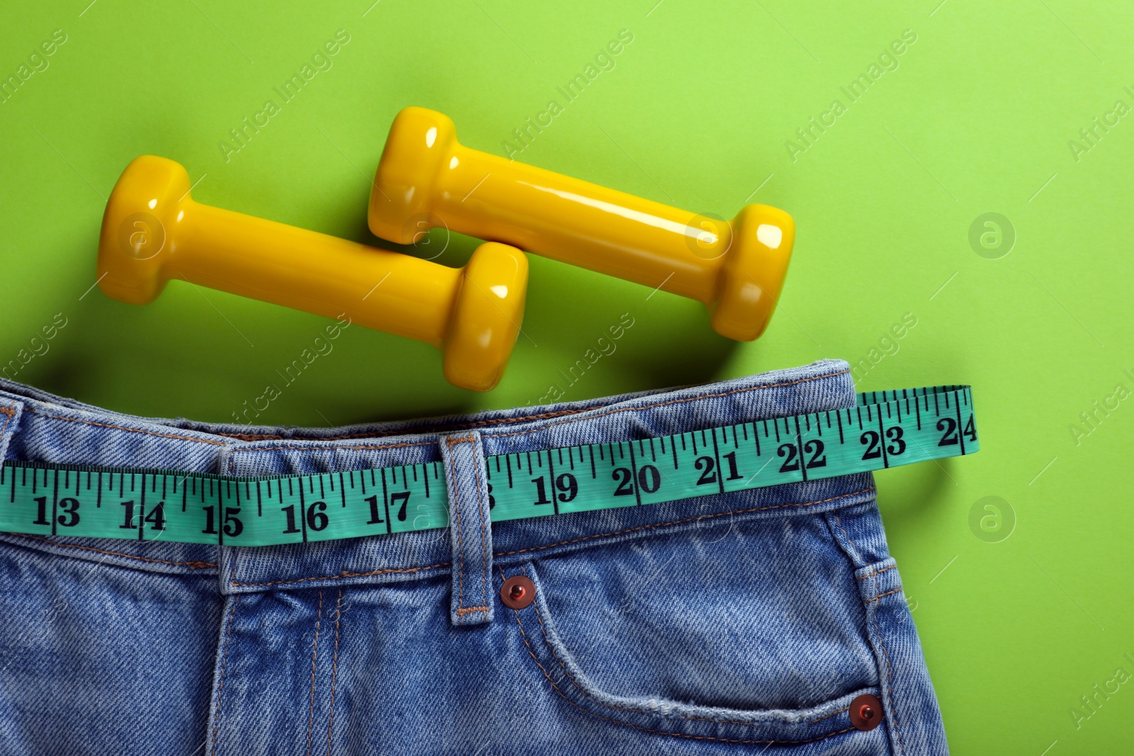 Photo of Jeans, dumbbells and measuring tape on light green background, flat lay. Weight loss concept