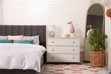 Photo of Comfortable bed with cushions, houseplant and chest of drawers in room. Stylish interior