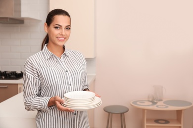 Photo of Beautiful young woman holding stack of clean dishes in kitchen