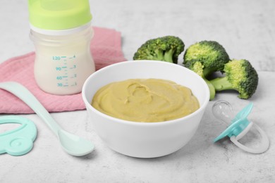 Bowl with healthy baby food, broccoli, spoon, pacifier and bottle of milk on white table, closeup