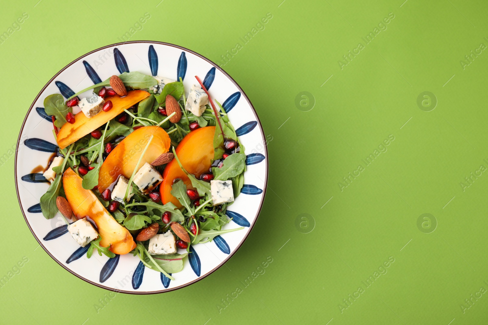 Photo of Tasty salad with persimmon, blue cheese, pomegranate and almonds served on light green background, top view. Space for text