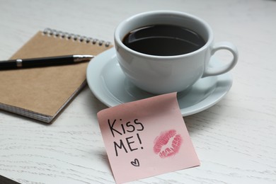 Photo of Sticky note with phrase Kiss Me, lipstick mark, cup of drink and notebook on white wooden table