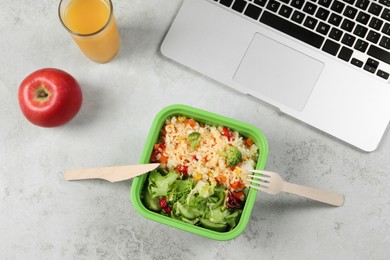 Container of tasty food, laptop, cutlery, apple and glass of juice on light grey table, flat lay. Business lunch