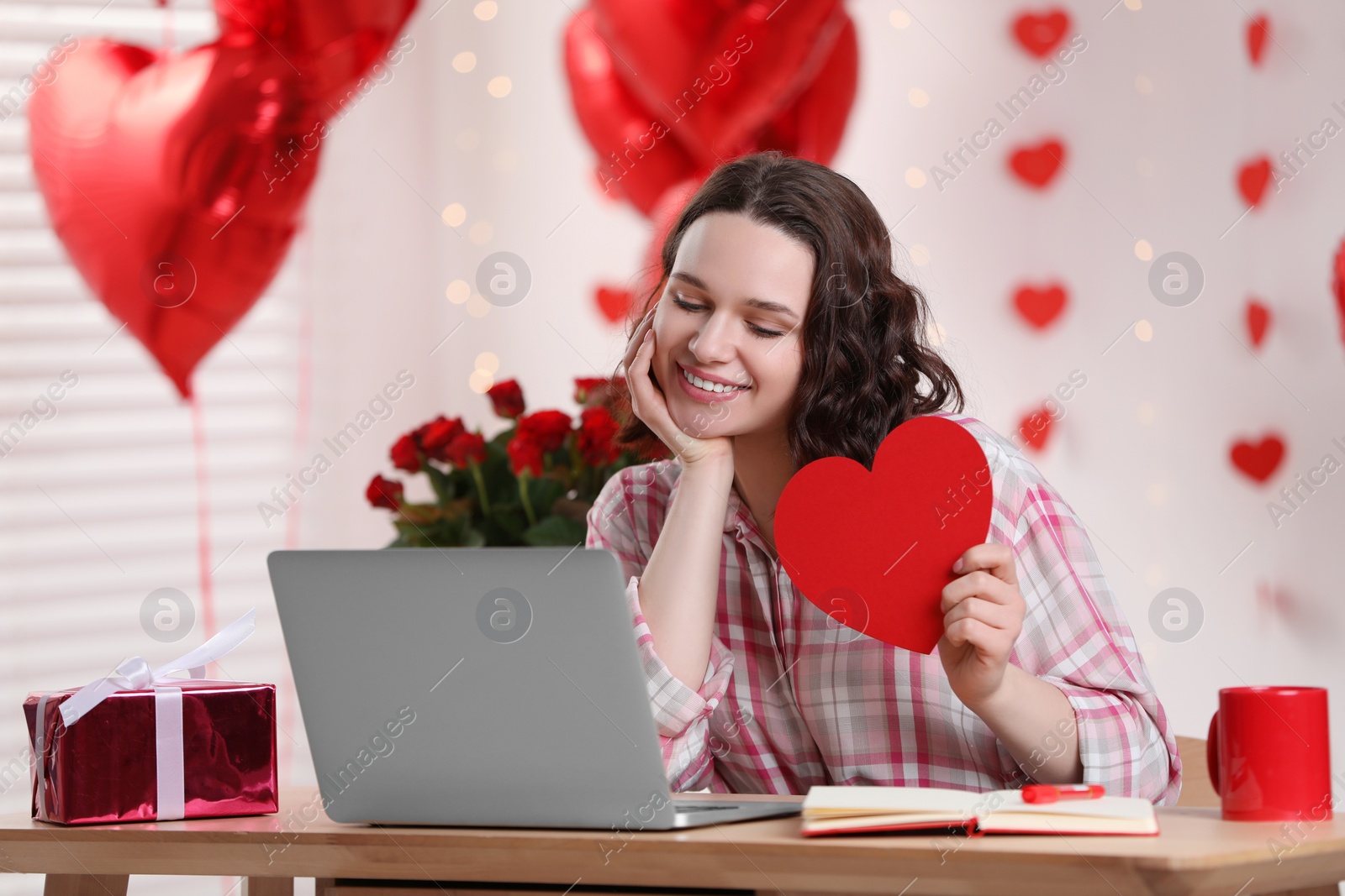 Photo of Valentine's day celebration in long distance relationship. Woman holding red paper heart while having video chat with her boyfriend via laptop at home