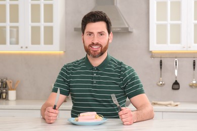 Photo of Happy man holding cutlery and plate of pasta with sausages at table in kitchen