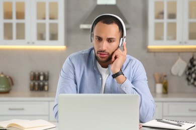 Photo of Young man with headphones working on laptop at desk in kitchen. Home office