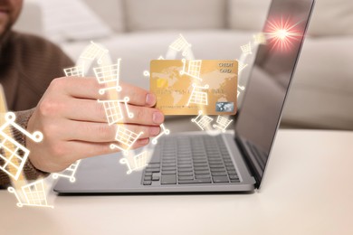 Man with credit card using laptop for online purchases at table, closeup. Illustrations of shopping cart around hand moving towards device screen