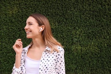 Young woman with wireless headphones listening to music near green grass wall. Space for text