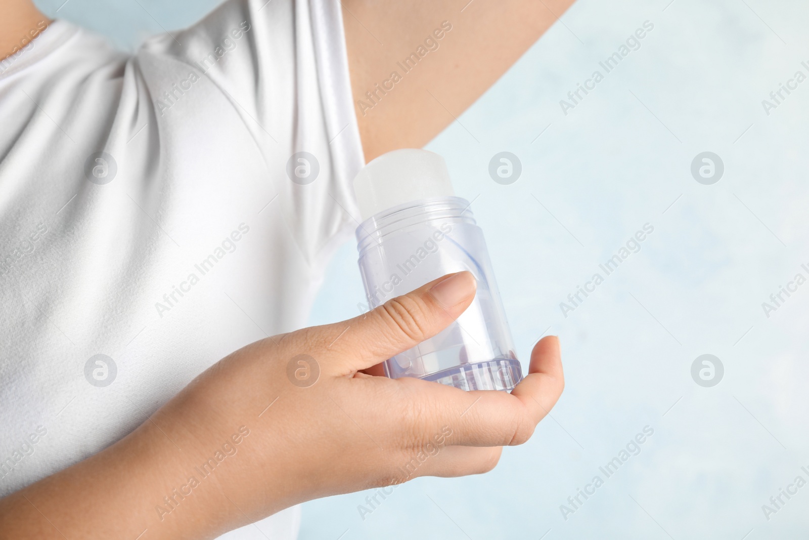 Photo of Young woman holding natural crystal alum deodorant near armpit on light blue background, closeup