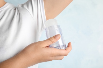 Photo of Young woman holding natural crystal alum deodorant near armpit on light blue background, closeup