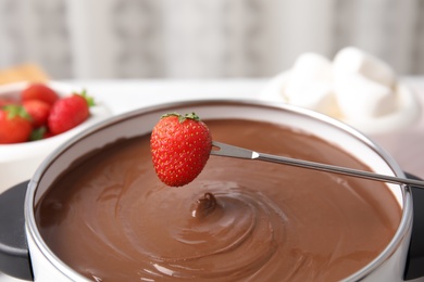 Dipping strawberry into pot with chocolate fondue on table, closeup