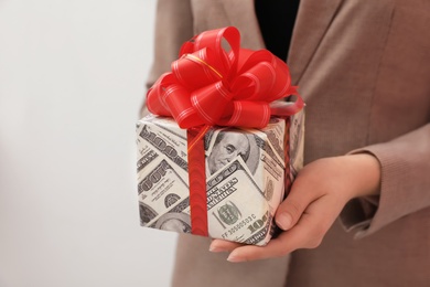 Woman holding gift box wrapped in dollars on light background, closeup