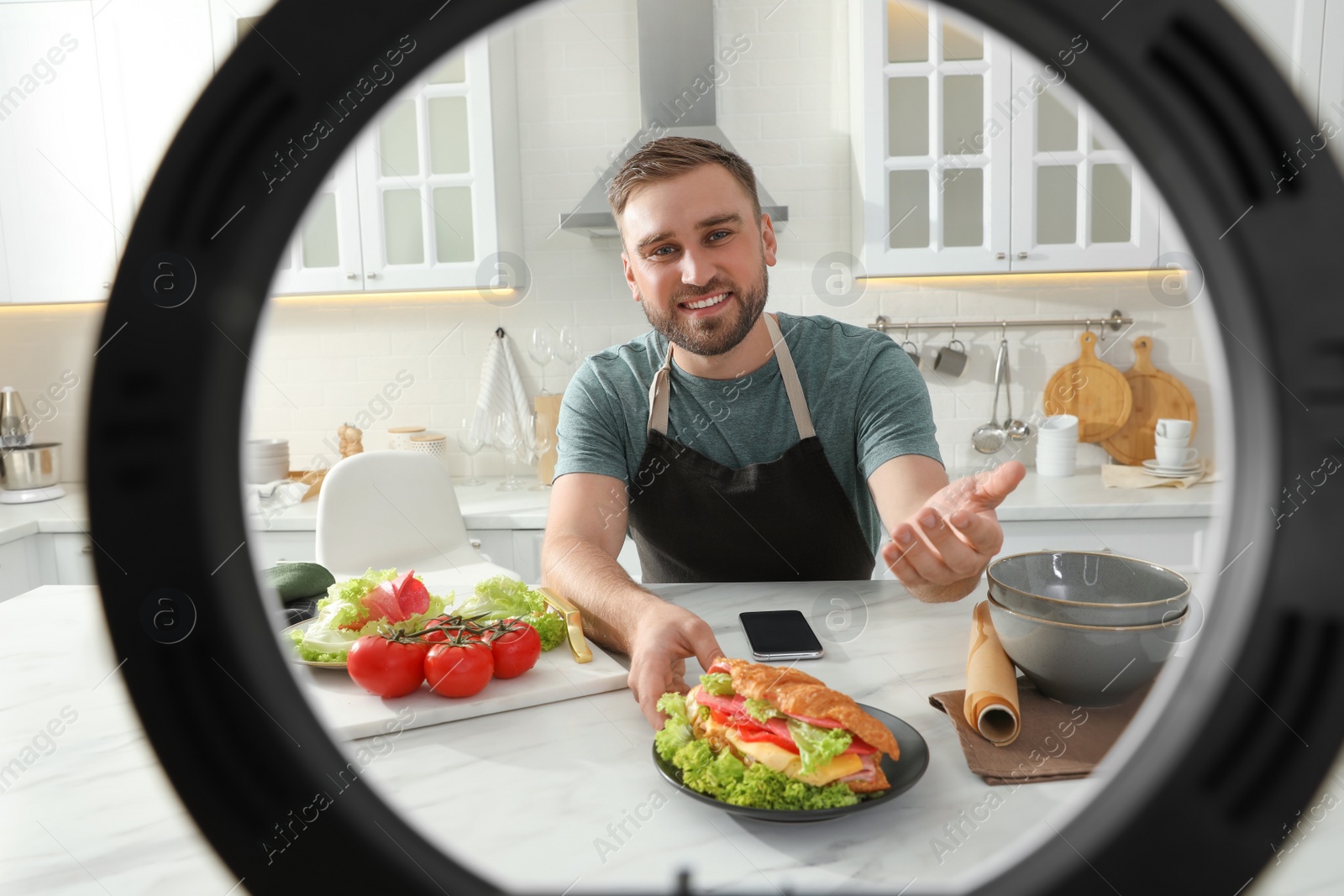 Photo of Blogger with tasty croissant sandwich recording video in kitchen at home, view through ring lamp