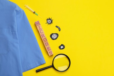 Photo of Word Microbes made with wooden cubes, magnifying glass, syringe and uniform on yellow background, flat lay. Space for text